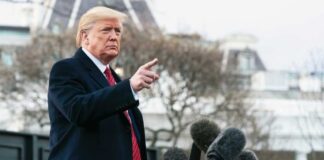 Man speaking to microphones, pointing outdoors.