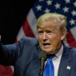 Trump Pardons Sheriff ‘Dragged Through Hell’ Man pointing while speaking at podium with American flags.