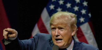 Man pointing while speaking at podium with American flags.