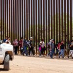 People walking along a border fence with patrol vehicle.