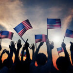 Silhouetted crowd holding American flags against a sunset
