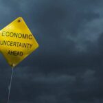 A yellow warning sign stating 'ECONOMIC UNCERTAINTY AHEAD' against a dark cloudy sky