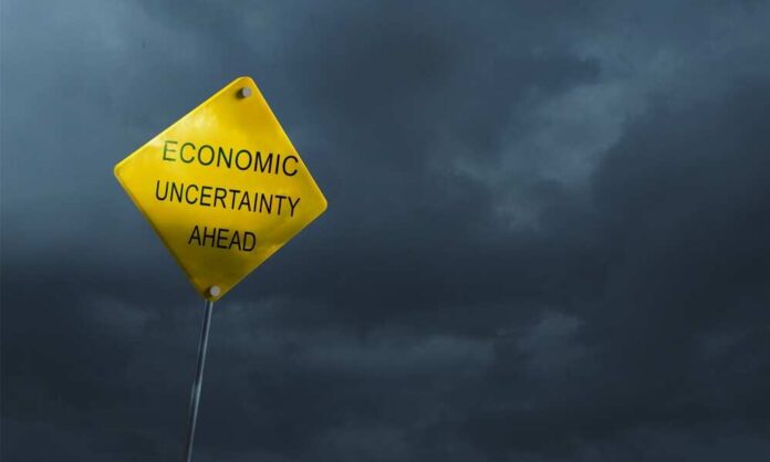 A yellow warning sign stating 'ECONOMIC UNCERTAINTY AHEAD' against a dark cloudy sky