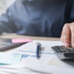 Person using a calculator while working on a laptop and reviewing documents