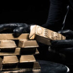 A person in black gloves handling gold bars against a dark background