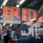 A traveler holding a suitcase in an airport terminal with a flight information display in the background