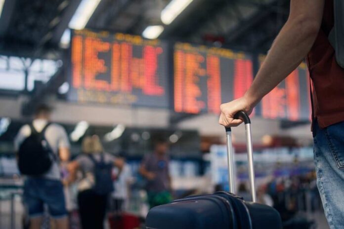 A traveler holding a suitcase in an airport terminal with a flight information display in the background