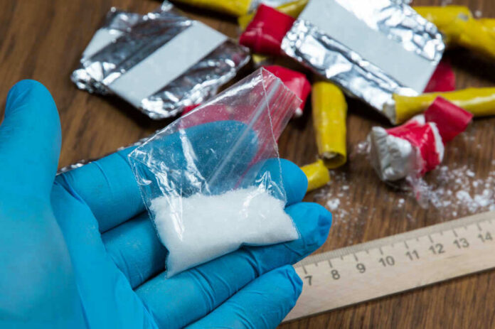 shutterstock_1955608786.jpg Blue-gloved hand holding bag of white powder, other substances nearby.