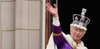 Man in royal attire and crown waving, stone background.