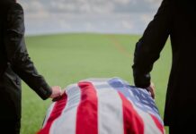 Two individuals standing beside a flag-draped casket in a green field