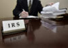 IRS sign on desk, person handling paperwork behind.