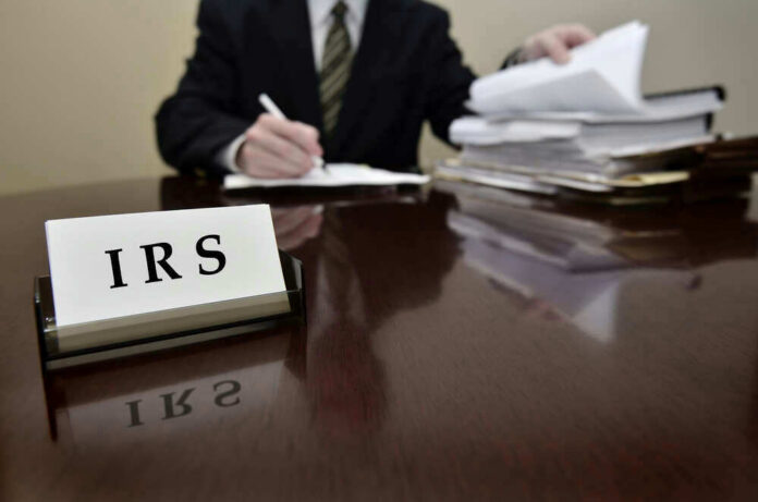 IRS sign on desk, person handling paperwork behind.