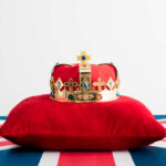 A royal crown resting on a red velvet pillow with a British flag background
