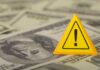 A yellow warning sign placed on a background of dollar bills