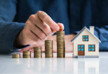 Hand stacking coins beside a miniature house model.