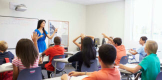 Teacher instructing students raising hands in a classroom.