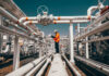 War Jolts Oil — $100 Whisper Grows Worker in orange jacket inspecting industrial pipes.