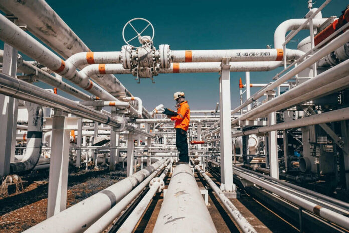 shutterstock_2420518787 Worker in orange jacket inspecting industrial pipes.