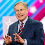 Extradition Standoff: Governor Abbott Holds The Clock Greg Abbott in suit speaking at an event.