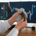Wall Street Euphoria — What’s Really Driving It? Person holding cash in front of stock market charts on computer screens