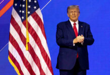 CEO Praise Of Trump Stuns D.C. Libs Donald Trump in suit near the American flag on stage.
