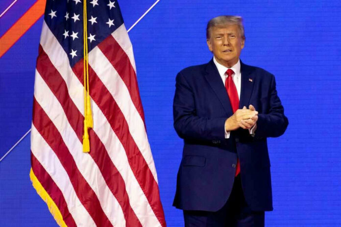 shutterstock_2272007717 Donald Trump in suit near the American flag on stage.