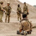 Soldier seated in desert foreground watching four armed soldiers standing