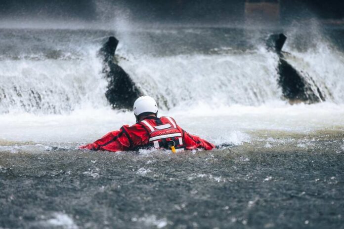 Firefighter,In,Water,Under,Dangerous,Wier,During,Drowning,Rescue,Training. Rescuer in red gear navigating turbulent water near a waterfall