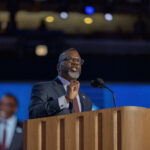 Governor Scrambles After Voter Bombshell Man in suit speaking at a podium with microphones
