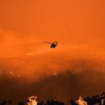 Helicopter flies over a wildfire at sunset with smoke and embers in the sky