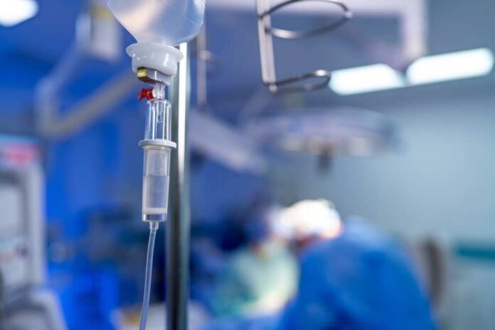 Close-up,Of,Iv,Drip,System,In,Hospital,Operating,Room.,Intravenous IV drip in focus with surgeons operating in the background