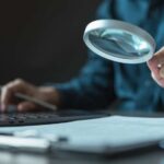 Person using a magnifying glass while working on a laptop