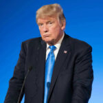 Man in suit with blue background speaking at podium.