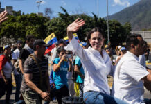 Chaos, Sky Shuts – Why Now? María Corina Machado waving in a street march alongside Teddy Williams