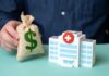 $50B ULTIMATUM Hits Hospitals Hand holding money bag beside toy hospital building