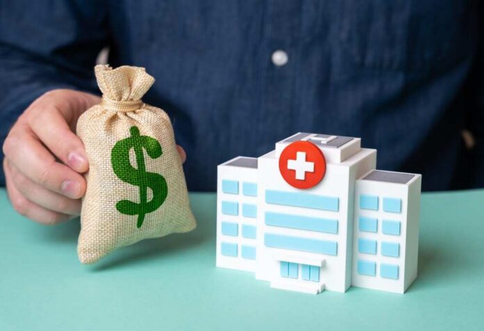 Hand holding money bag beside toy hospital building